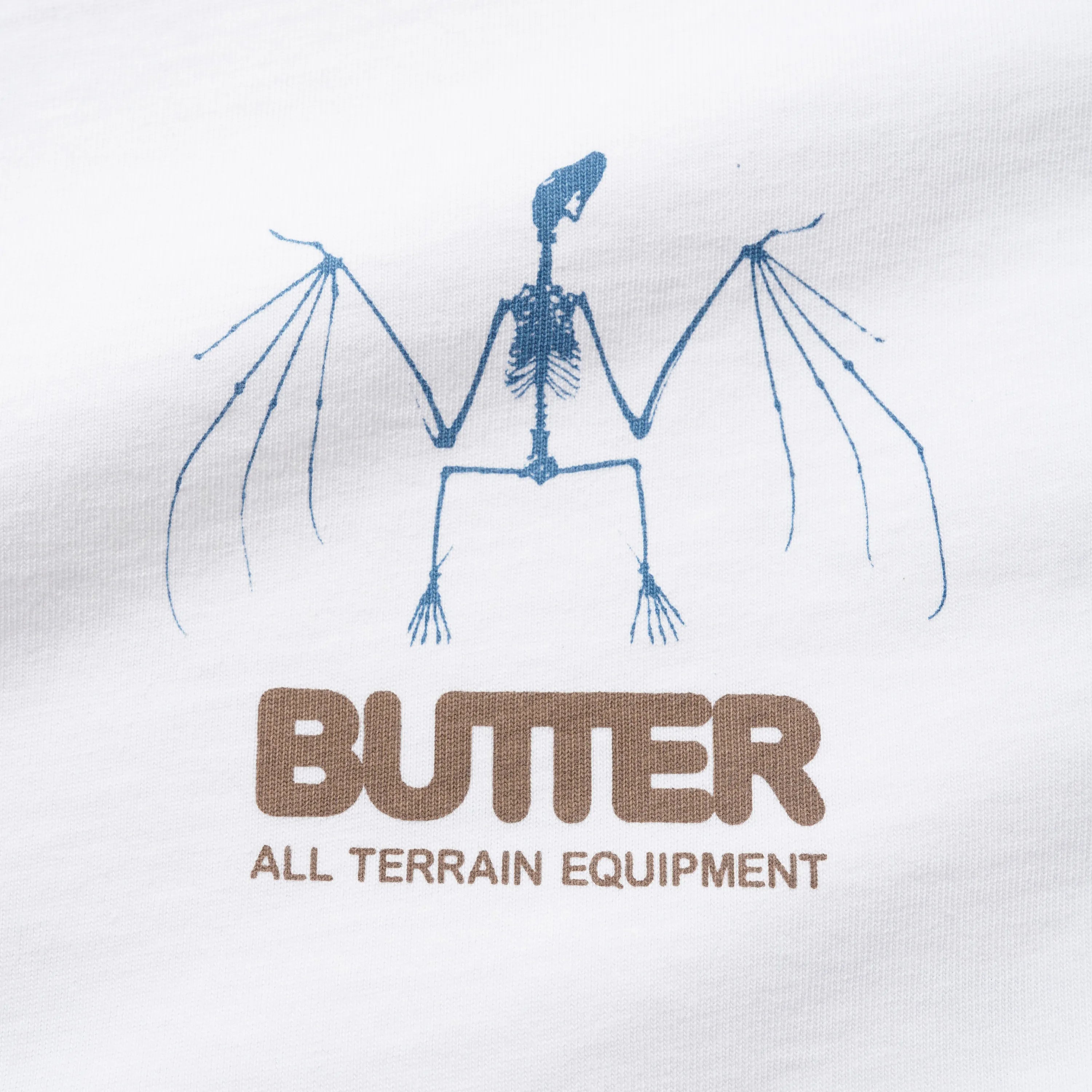 Gros plan sur la sérigraphie poitrine du t-shirt Butter Goods montrant un squelette bleu et le logo marron All Terrain Equipment.