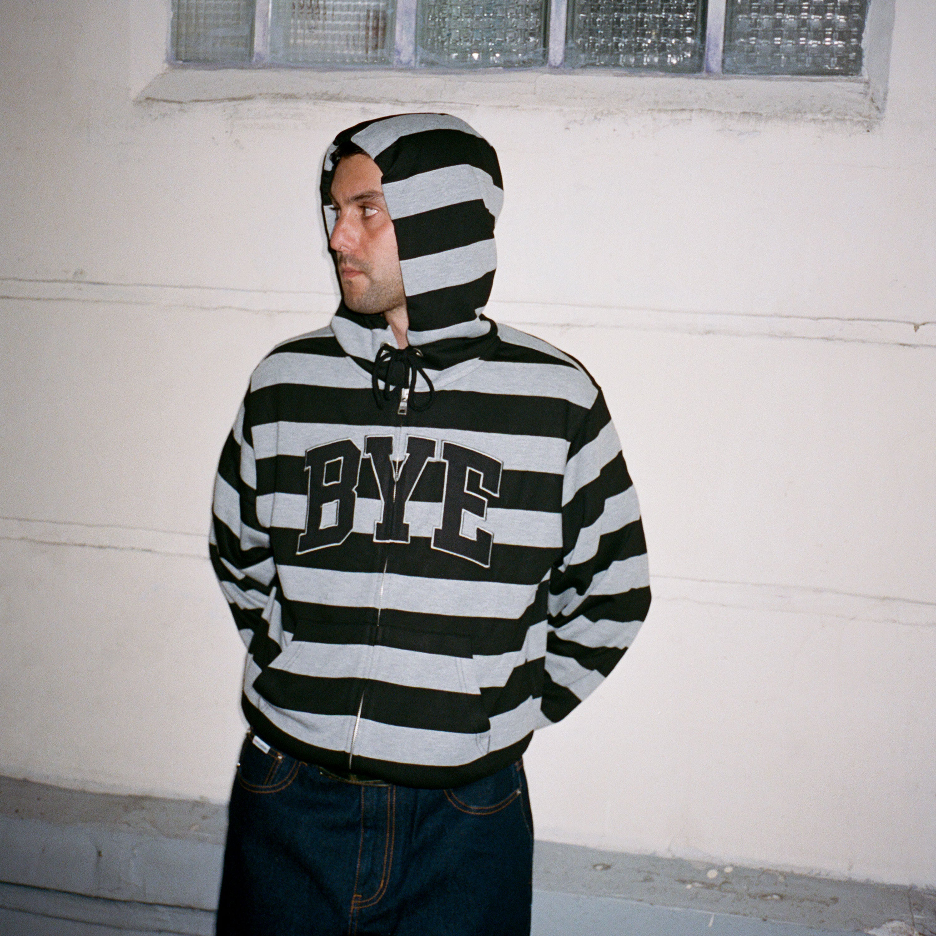 Photo lookbook sweat à capuche zippé Bye Jeremy modèle Bye Hoodie Stripes, avec des rayures horizontales épaisses grises et noires et un grand logo BYE .